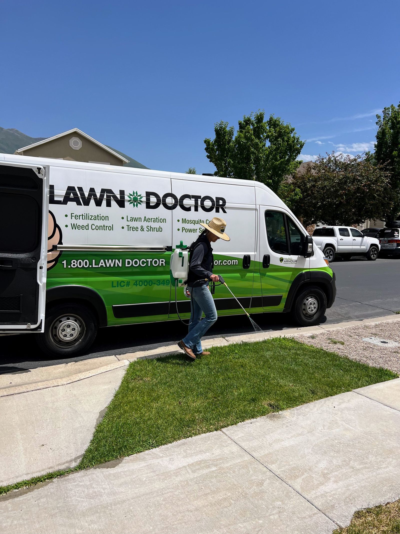 Lawn Doctor technician in a straw hat applying a treatment next to a green and white Lawn Doctor service vehicle.
