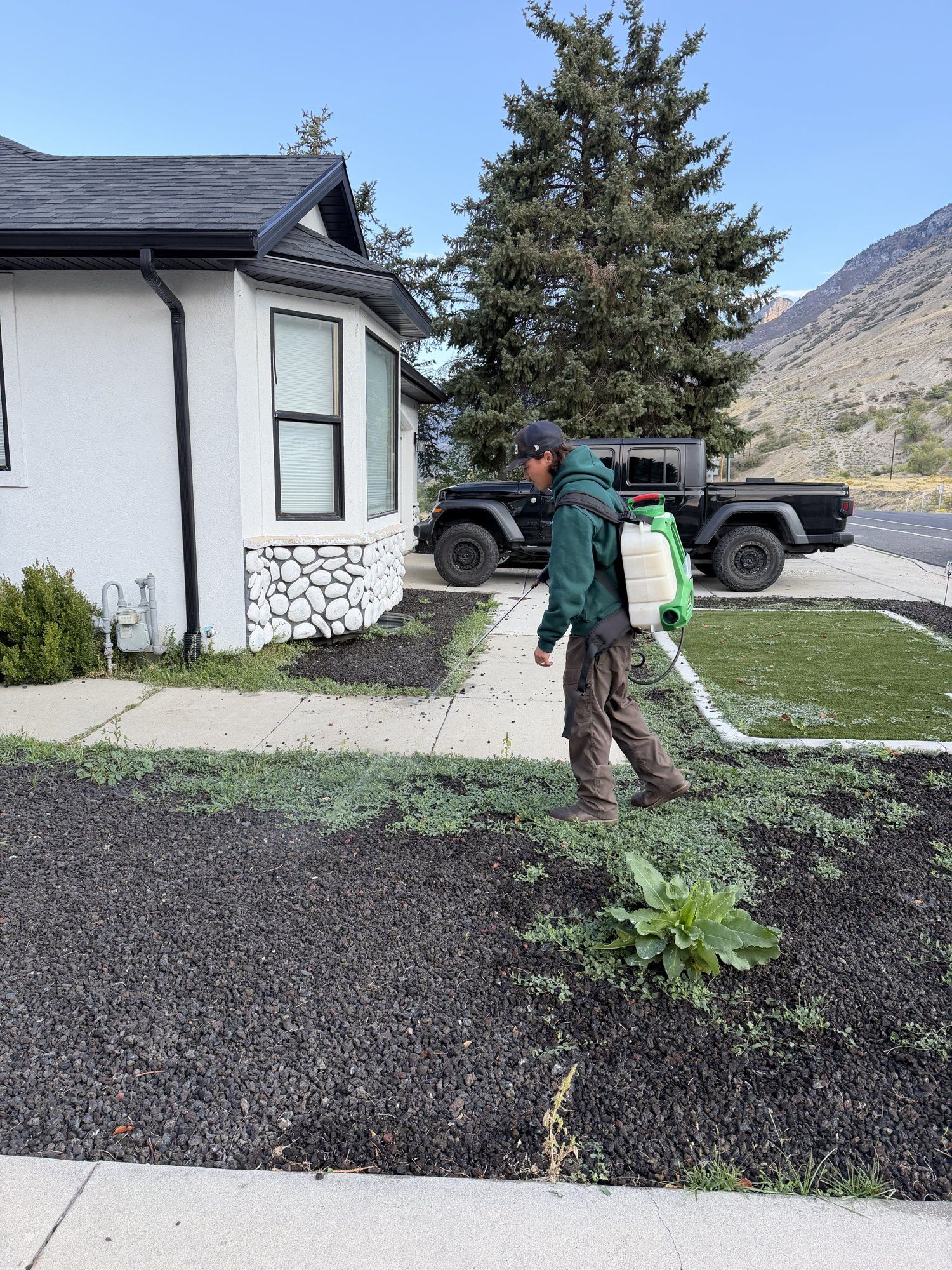 Lawn Doctor technician with a backpack sprayer treating weeds growing through a lava rock bed beside a sidewalk at a Utah home.